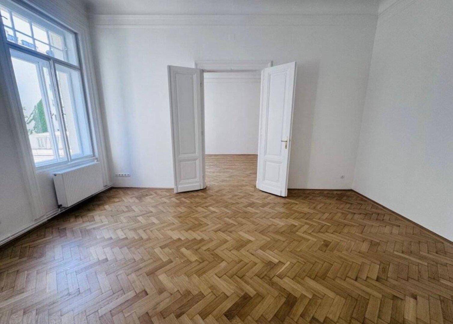 Geräumiges Zimmer mit Fischgrätparkett, hohen Decken und eleganten Doppeltüren.