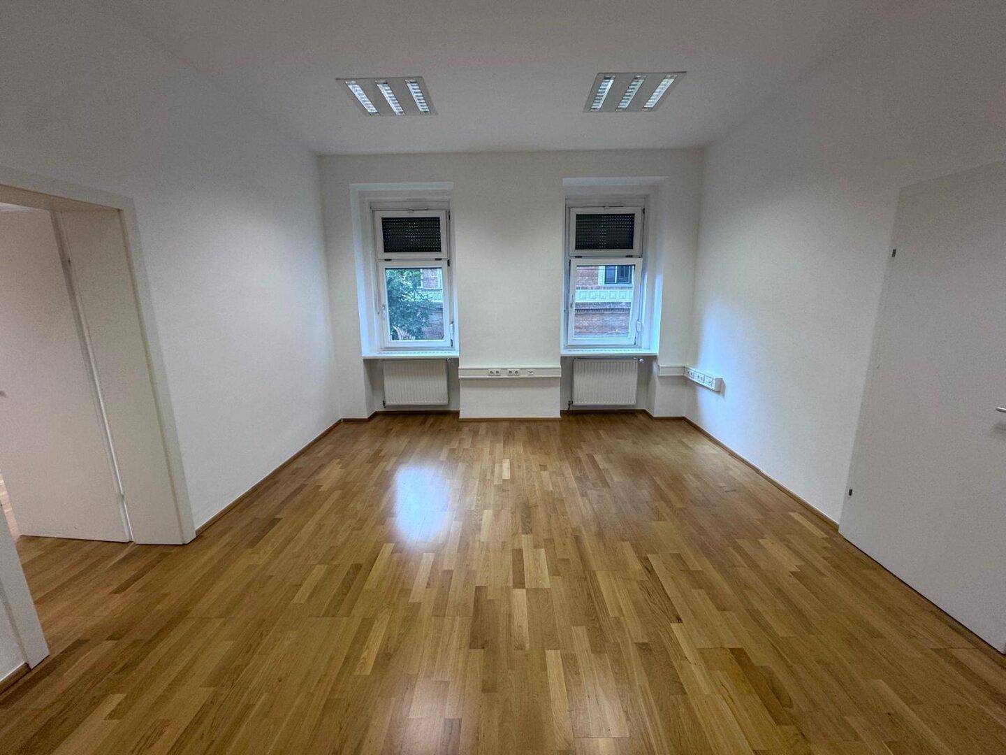 Heller Büroraum mit zwei Fenstern, Holzparkett und funktionaler Deckenbeleuchtung.