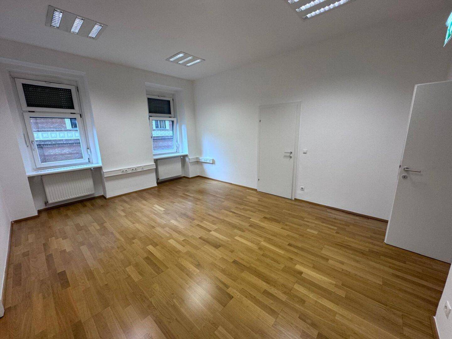 Leerer Büroraum mit Holzparkett, zwei Fenstern und weiß gestrichenen Wänden, bereit zur Nutzung.