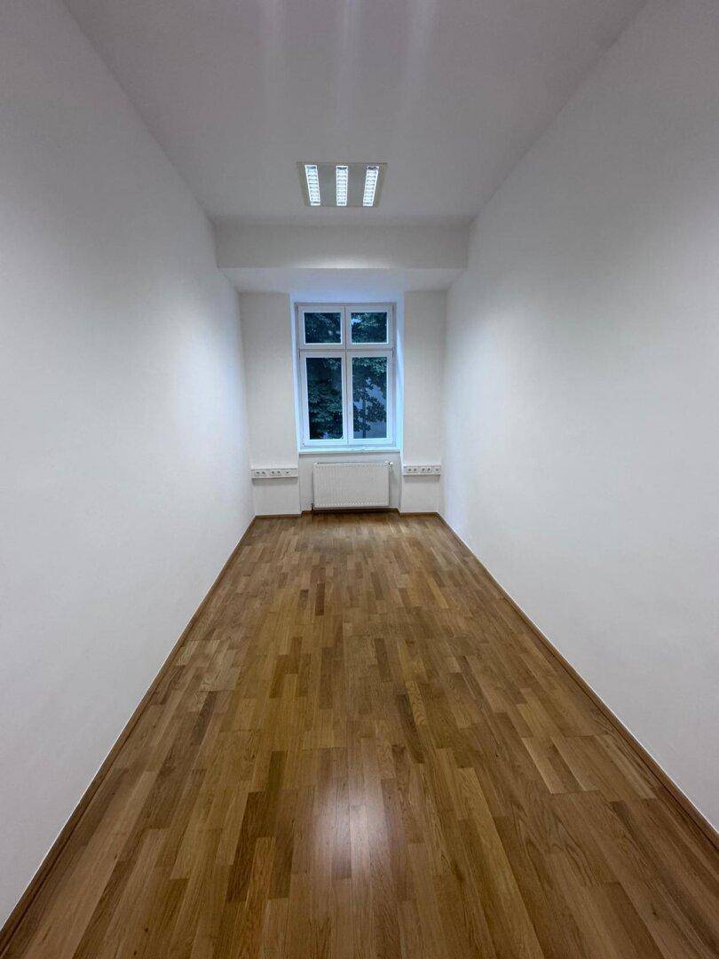 Langer, schmaler Raum mit einem Fenster und hellem Holzparkett, ideal als Einzelbüro.