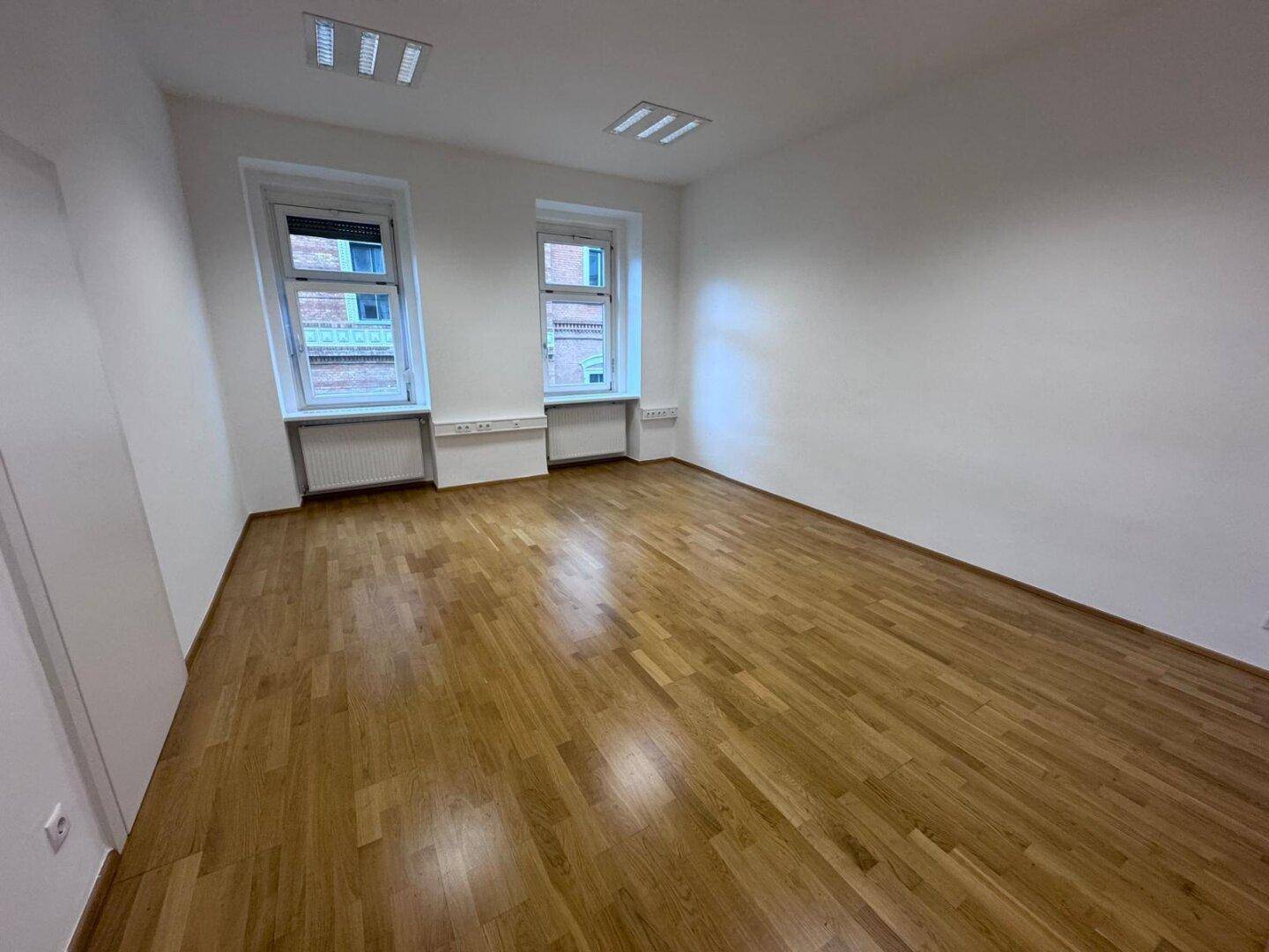 Leerer Büroraum mit Holzparkett, zwei Fenstern und weiß gestrichenen Wänden, bereit zur Nutzung.