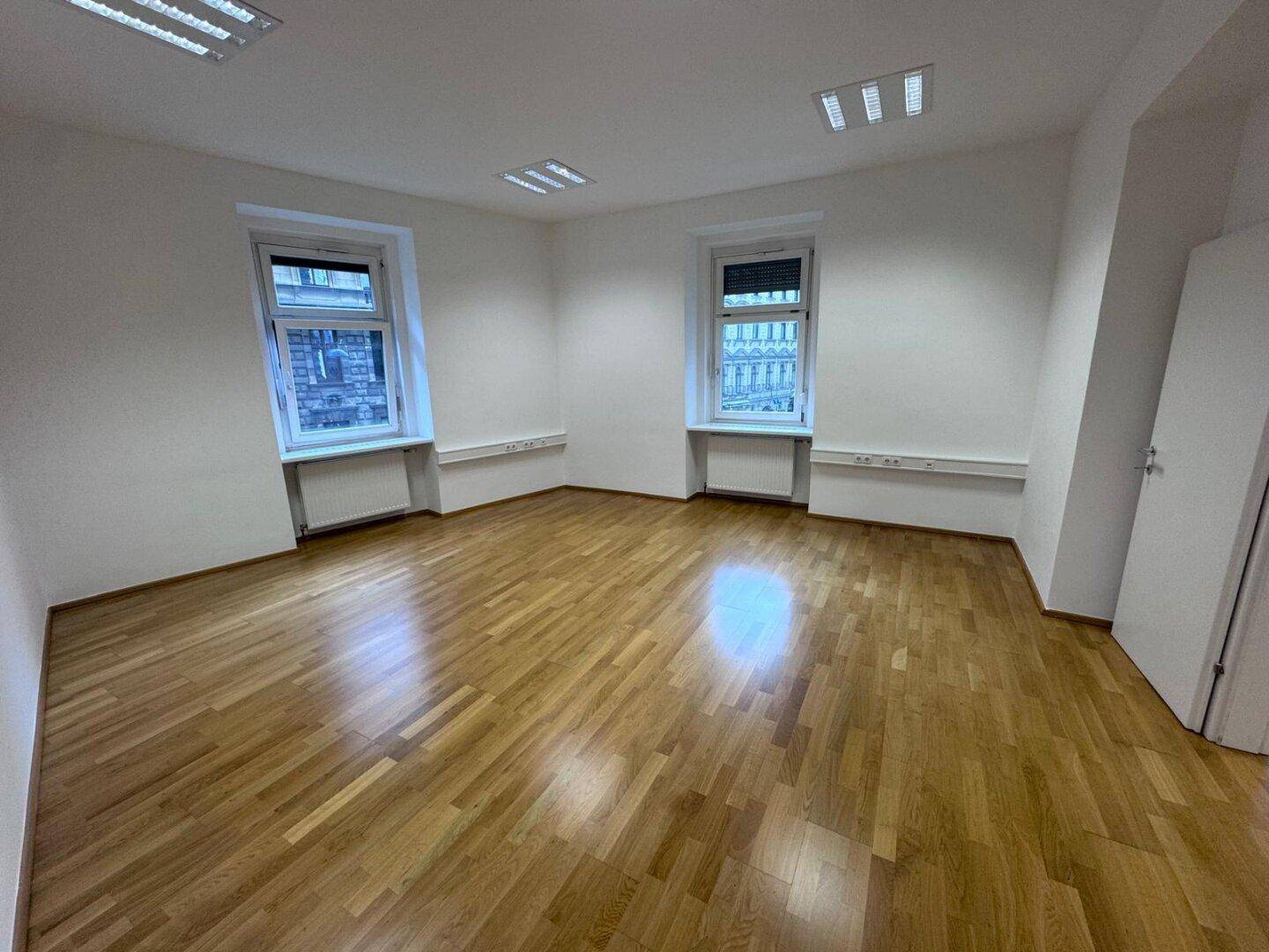 Heller Büroraum mit zwei Fenstern, Holzparkett und weiß gestrichenen Wänden.