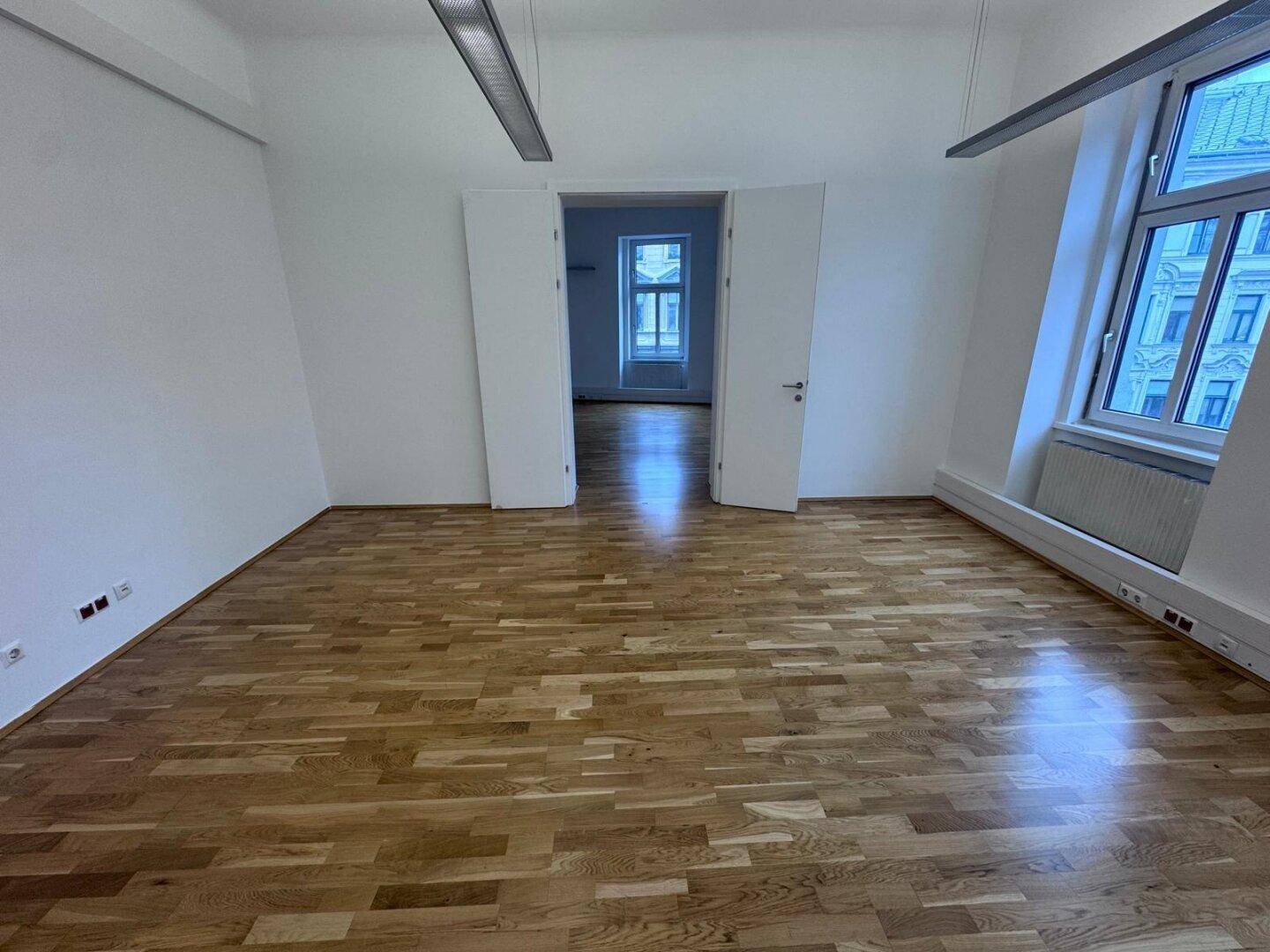 Geräumiger Büroraum mit Holzboden und Blick durch eine Doppeltür in einen weiteren hellen Bereich.