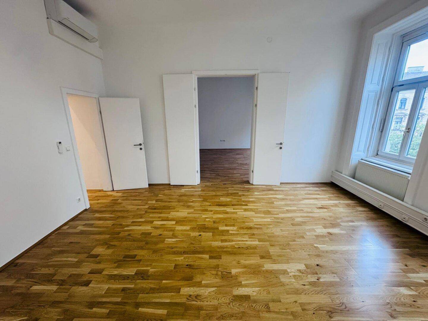 Heller Büroraum mit Holzboden, Klimaanlage und offener Doppeltür zu einem angrenzenden Bereich.