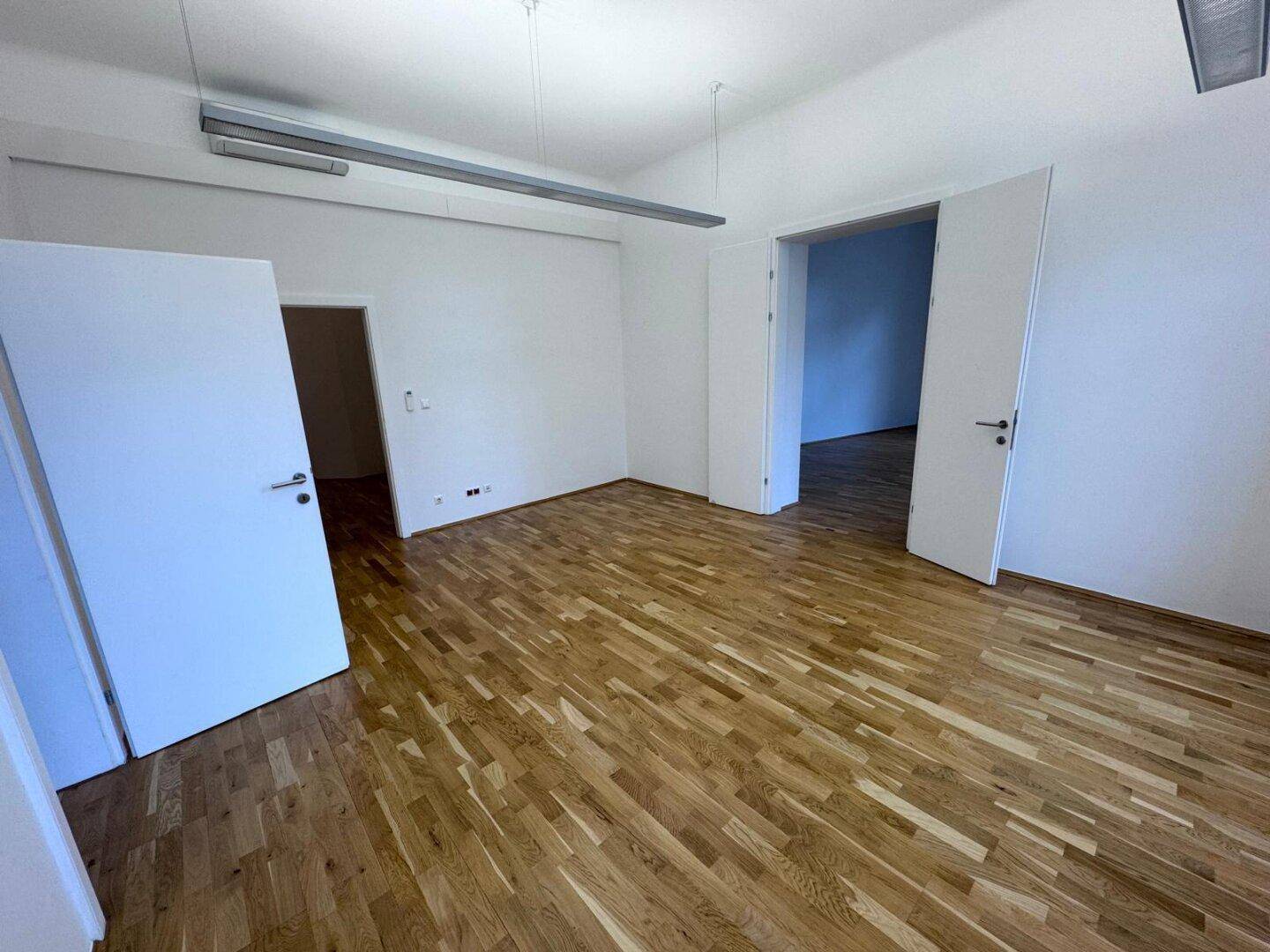 Geräumiger Büroraum mit Holzboden, weißen Wänden und offener Doppeltür zu einem Nebenraum, vielseitig nutzbar.