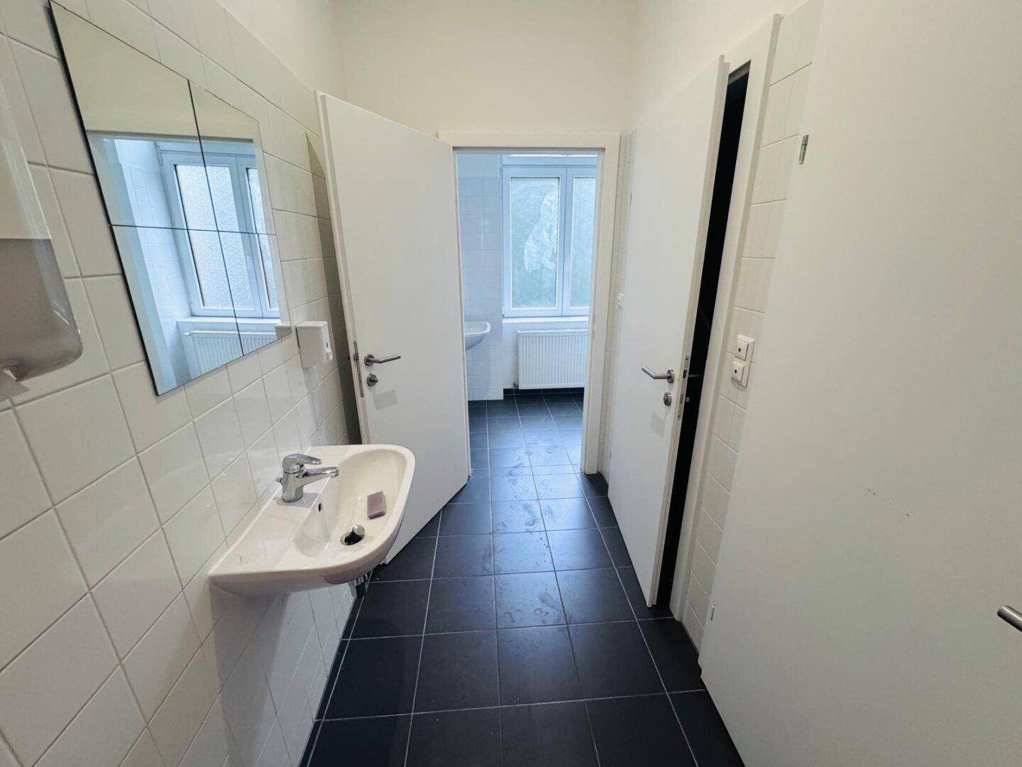 Schlichtes Badezimmer mit weißen Fliesen, Waschbecken und einem Fenster für natürliche Belüftung und Tageslicht.
