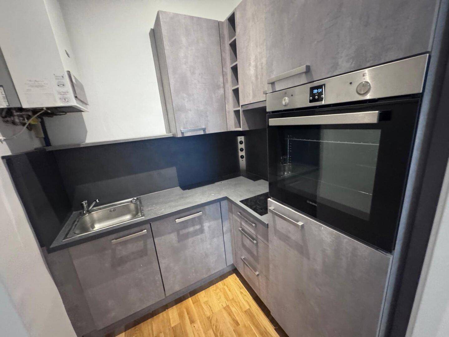 Kompakte Küchenzeile mit grauen Schränken, integriertem Backofen und Spüle, ideal für kleine Büros oder Apartments.