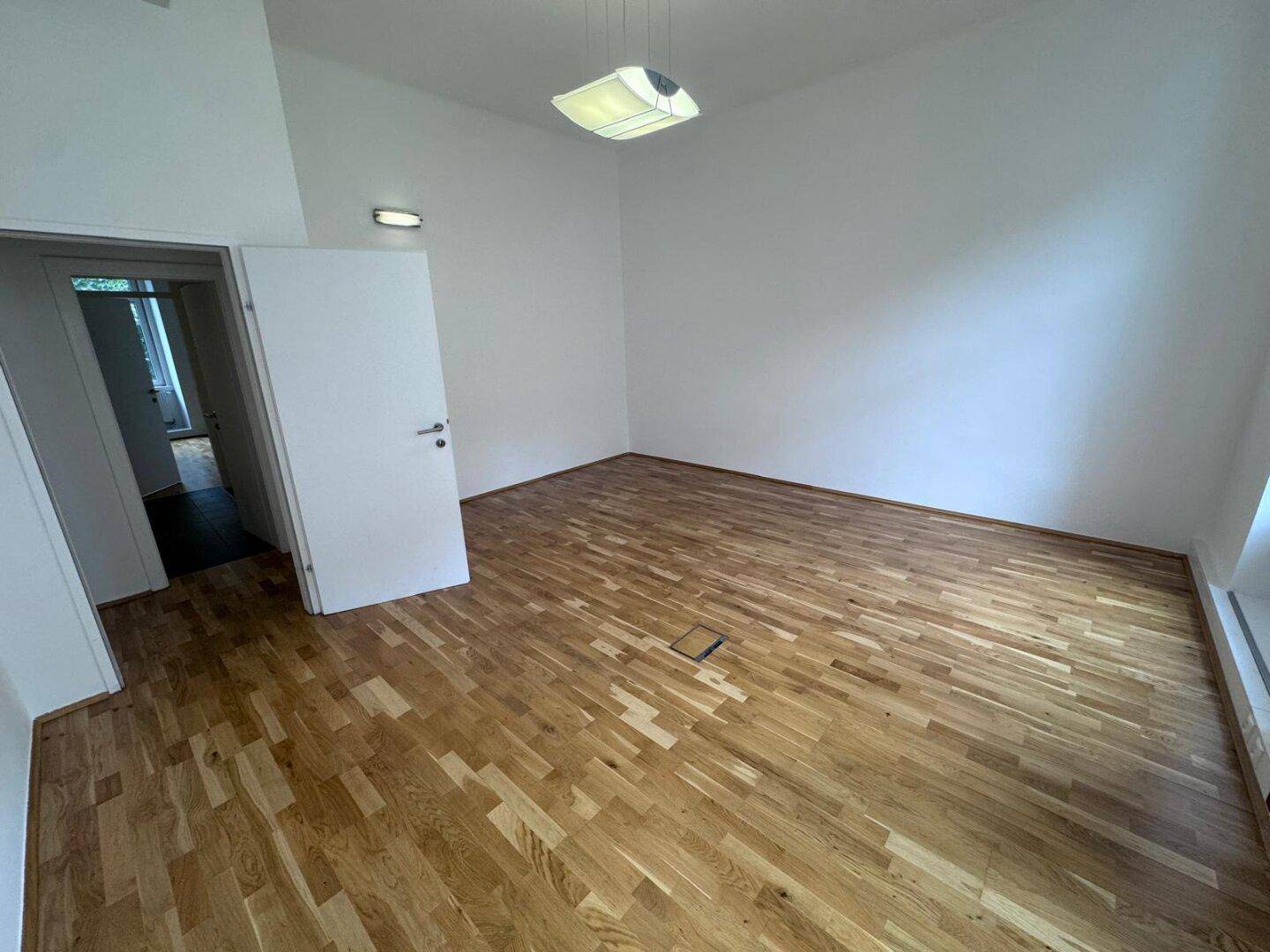 Heller Büroraum mit Holzboden, weißen Wänden und einer modernen Deckenleuchte, bietet eine angenehme Arbeitsatmosphäre.