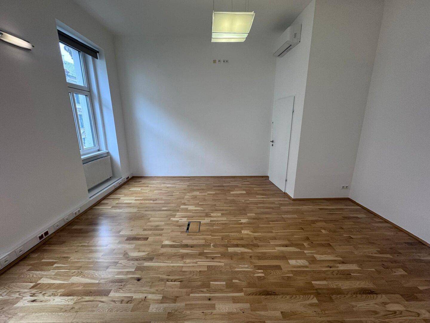 Heller Büroraum mit Holzboden, Klimaanlage und einem Fenster mit Blick auf die Straße, komfortabel und funktional.