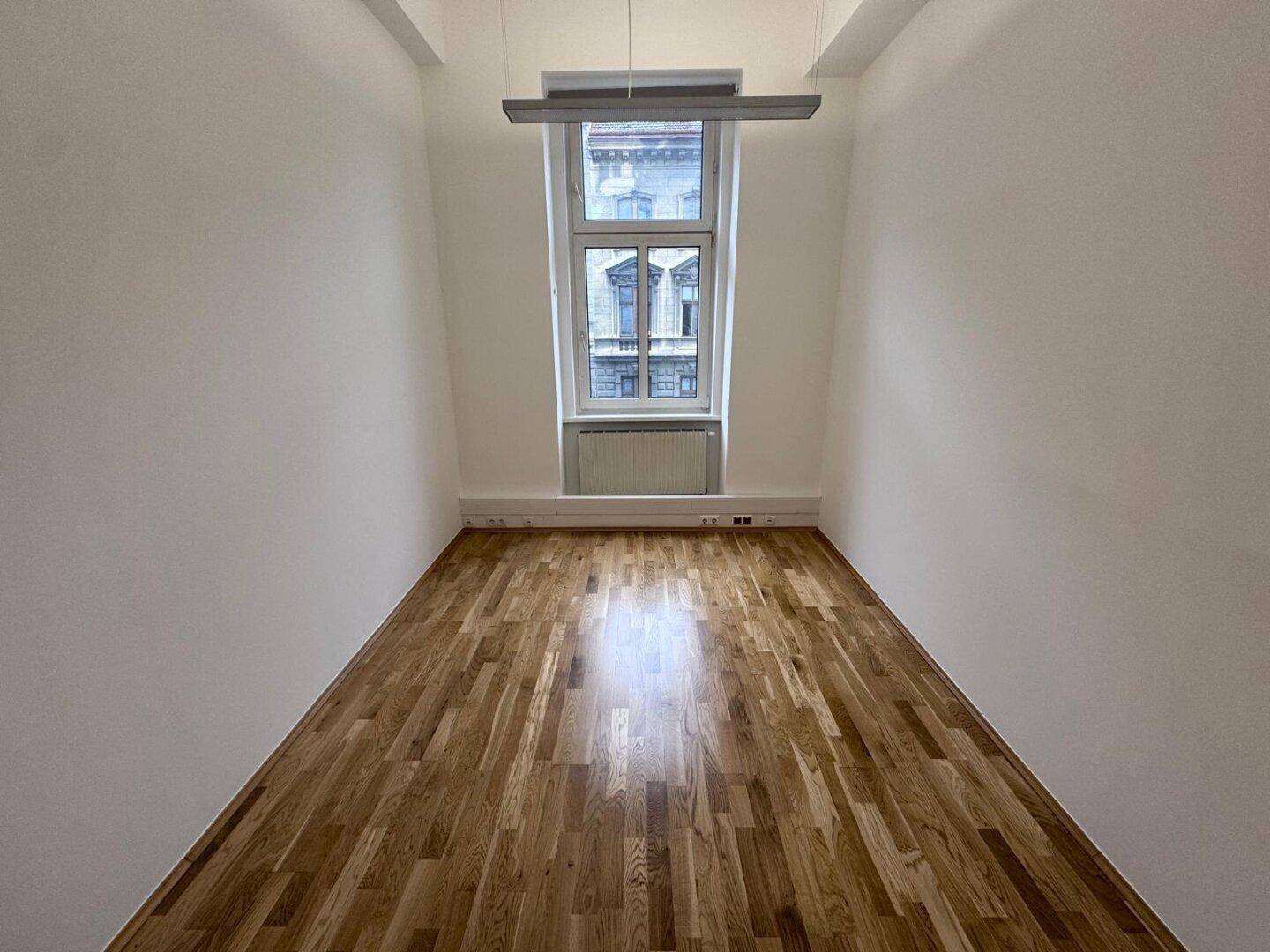 Heller Büroraum mit Holzboden, einem großen Fenster und einer modernen Deckenleuchte, bietet viel Tageslicht.