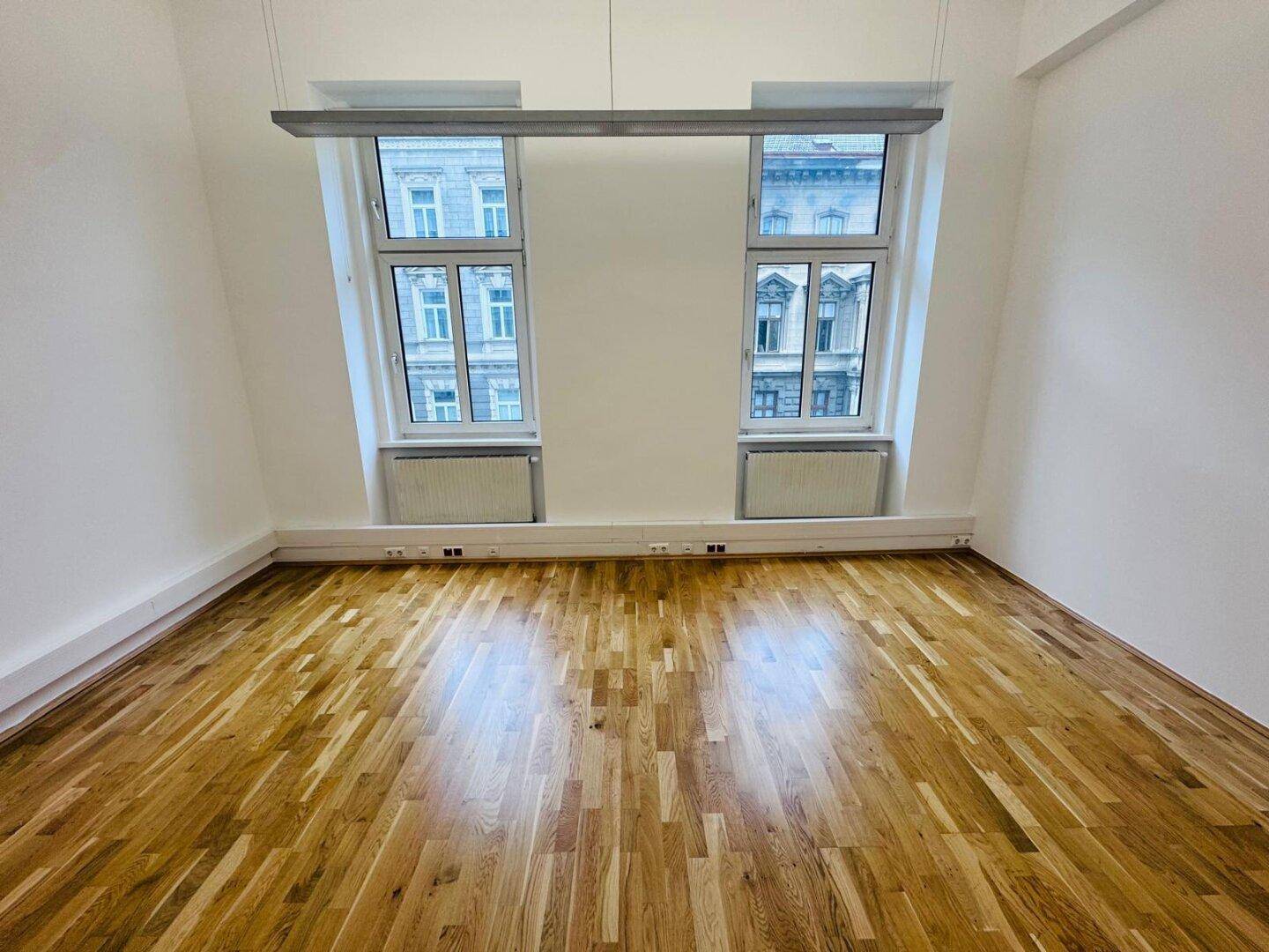 Heller Büroraum mit zwei Fenstern, Holzboden und Blick auf die umliegenden Gebäude, bietet viel Tageslicht.