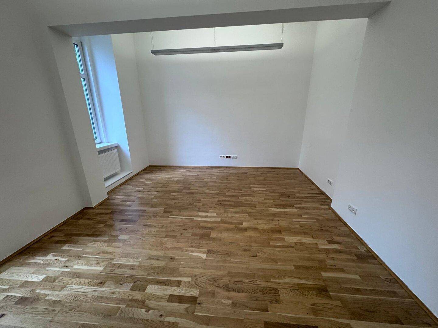 Heller Büroraum mit Holzboden, weißen Wänden und einer modernen Deckenleuchte, schafft eine angenehme Arbeitsumgebung.