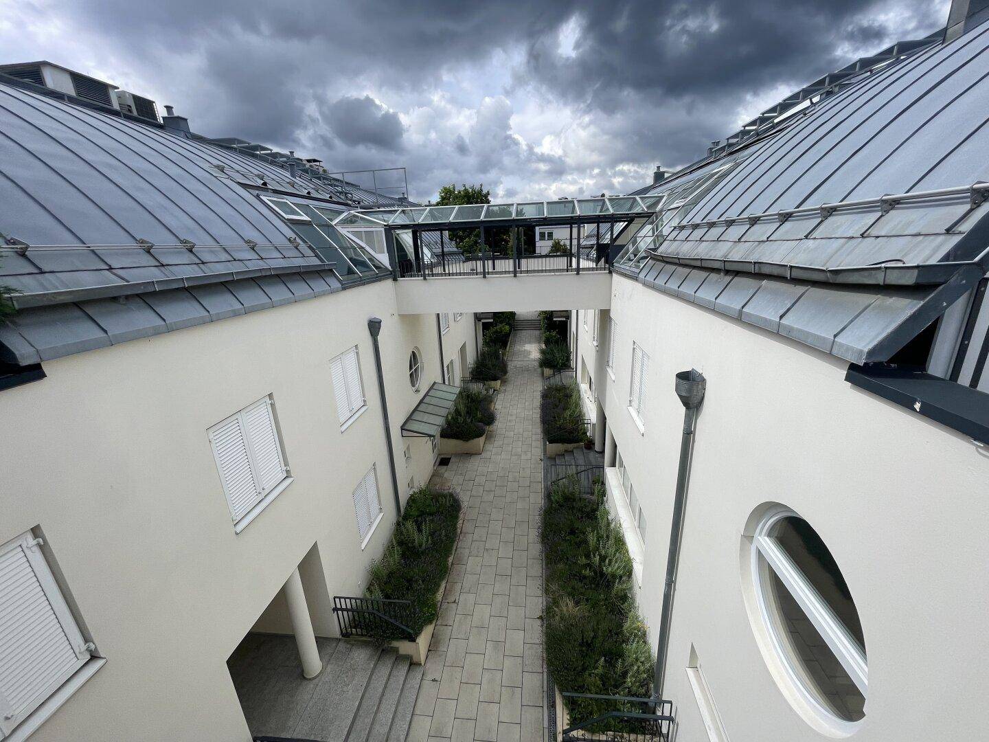 Innenhof zwischen Gebäuden mit gepflastertem Weg, Bepflanzung und einer Glasbrücke.