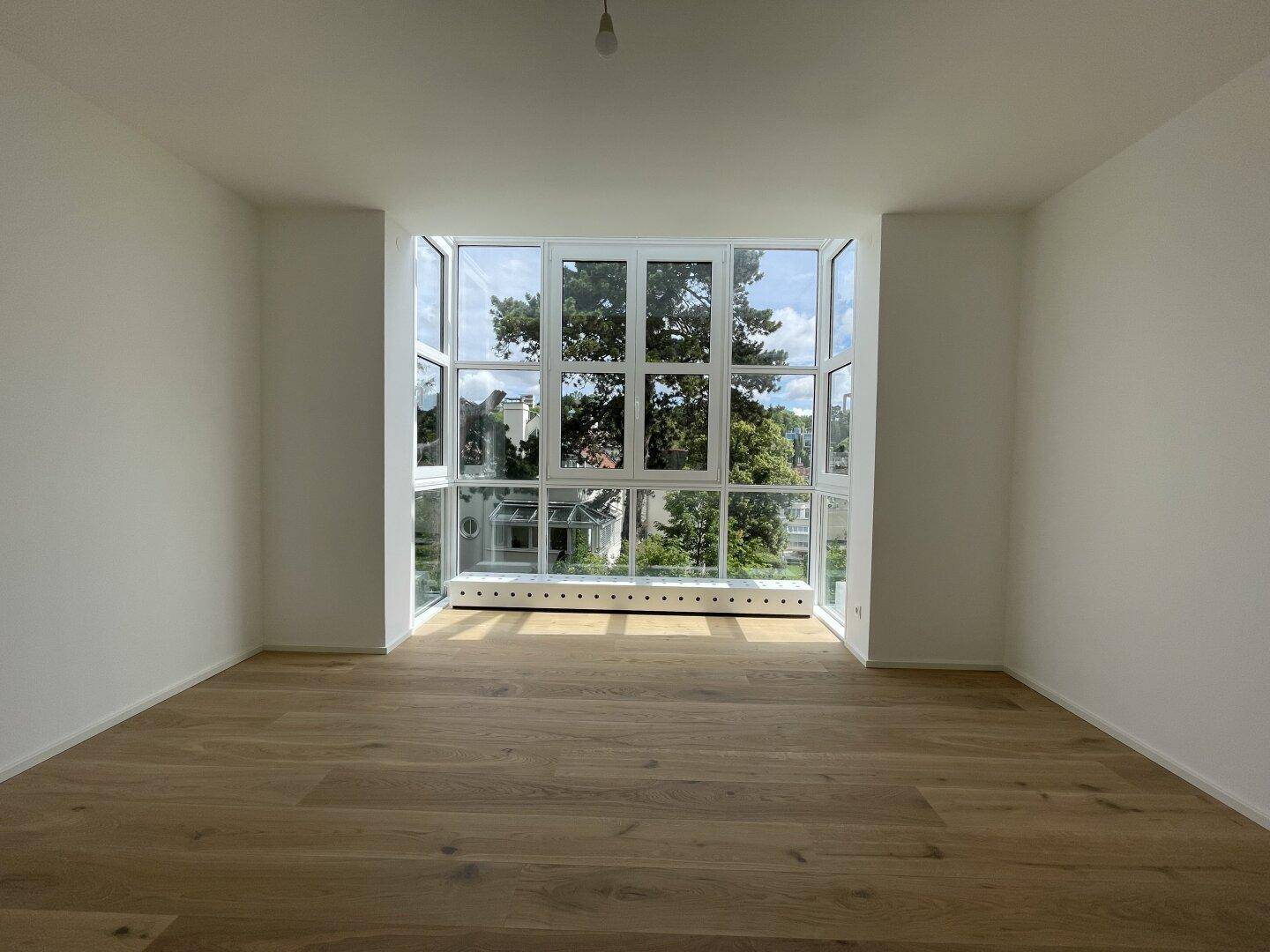 Helles Zimmer mit Holzboden und großen, bodentiefen Fenstern mit Blick ins Grüne.