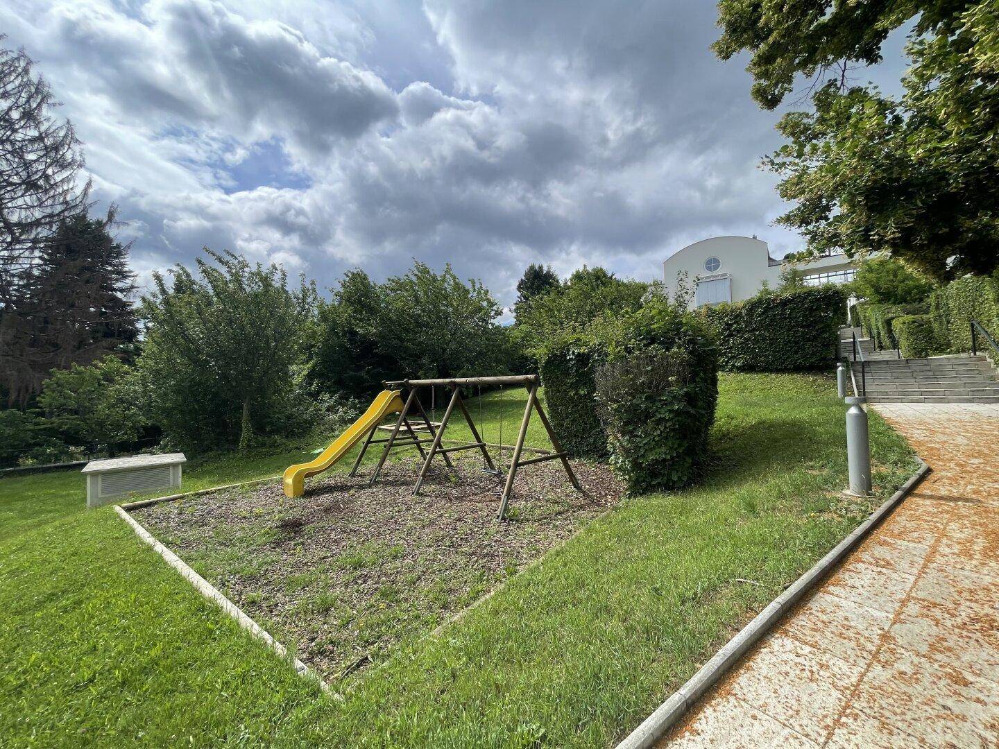 Kinderspielplatz im Freien mit Schaukel und Rutsche, umgeben von Grünflächen und Bäumen.