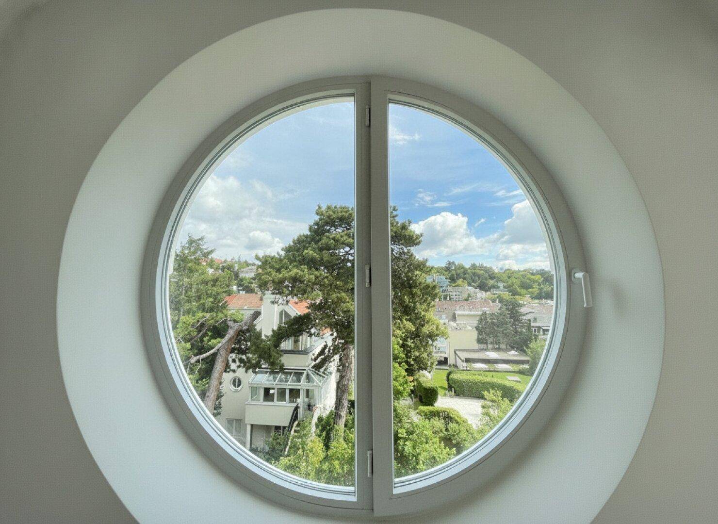 Rundes Fenster mit weitem Blick auf grüne Bäume und die umliegende Wohngegend.