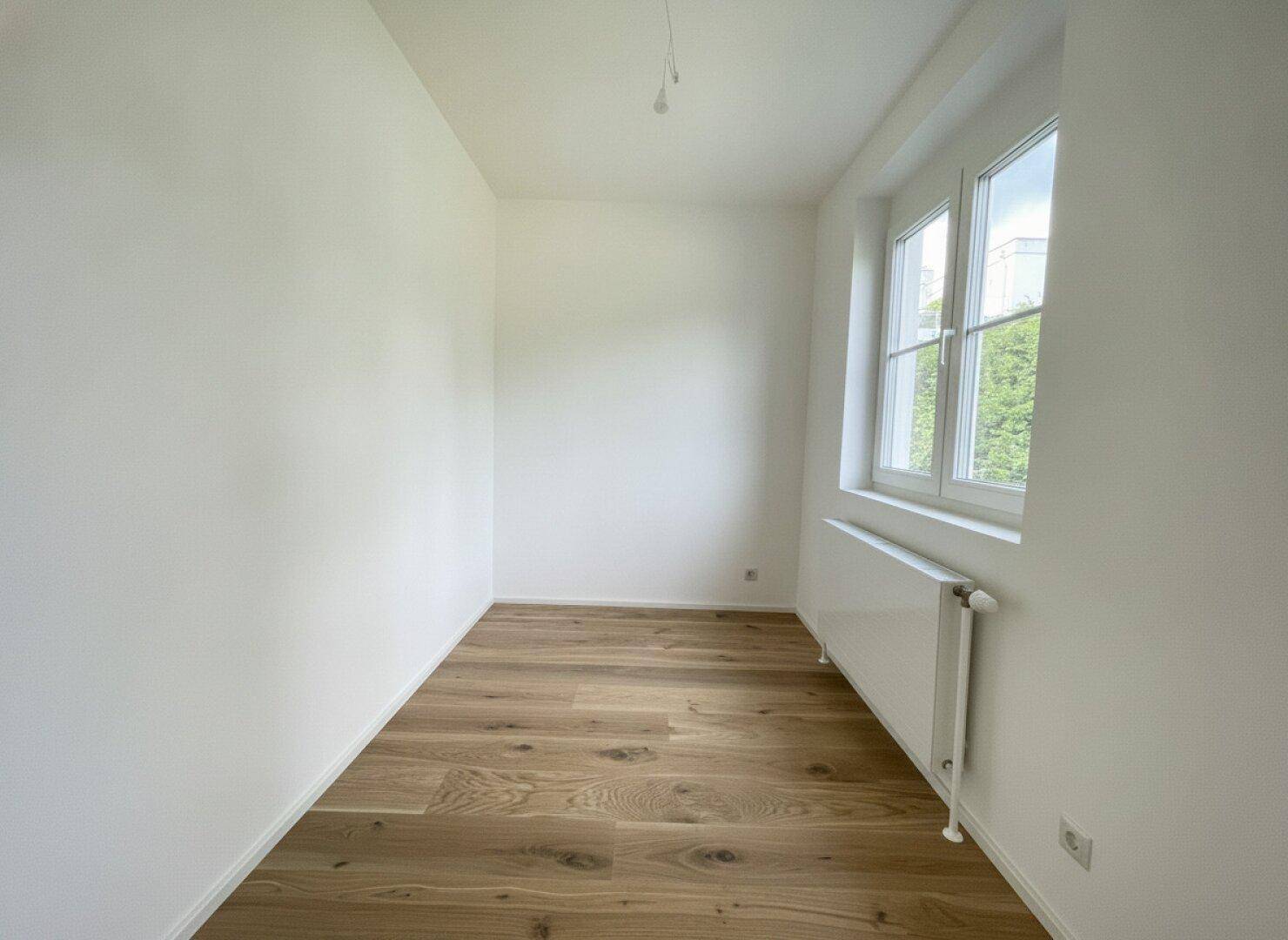 Ruhiges Zimmer mit Holzboden, weiß gestrichenen Wänden und einem Fenster mit Blick ins Grüne.