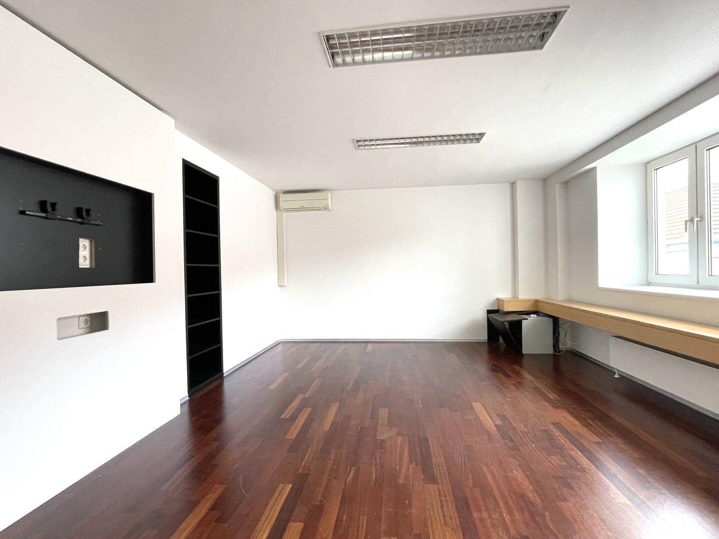 Helles Büro mit dunklem Holzboden, weißen Wänden und Klimaanlage über dem Fenster.
