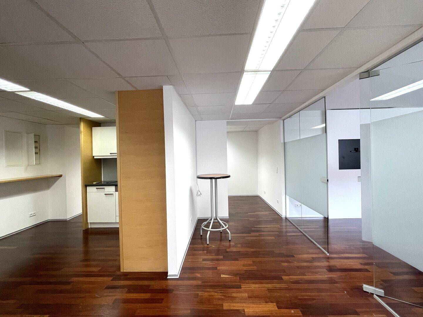 Geräumiges Büro mit dunklem Holzboden, weißen Wänden und einer kleinen Küchenzeile im Hintergrund.