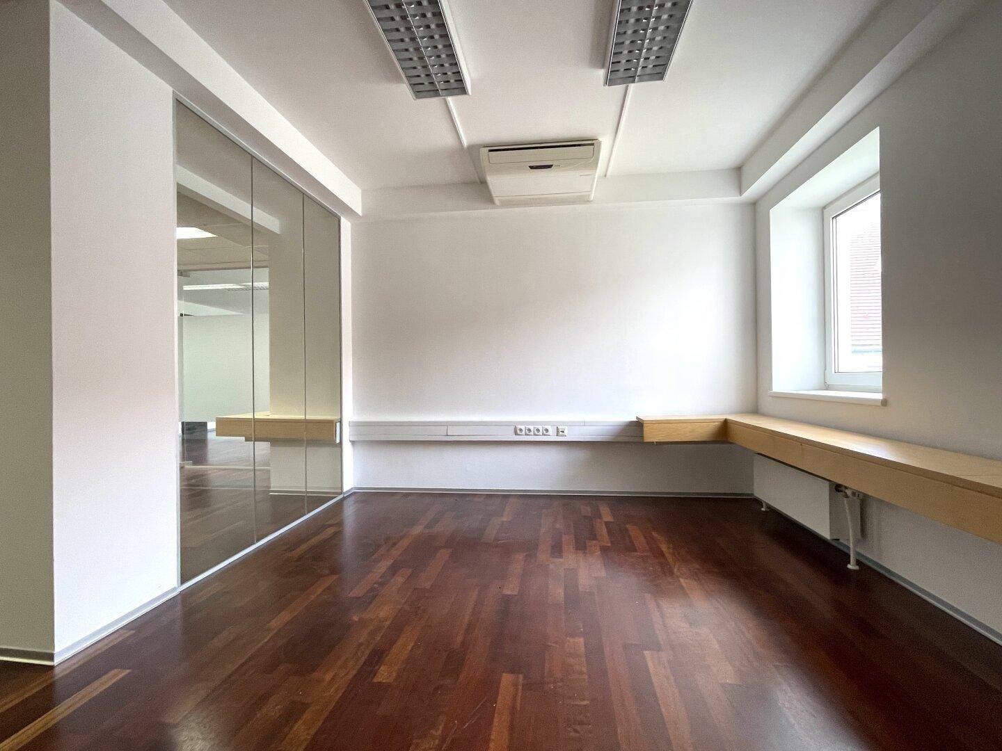 Büro mit dunklem Holzboden, Glaswänden und einer langen Arbeitsplatte unter dem Fenster.