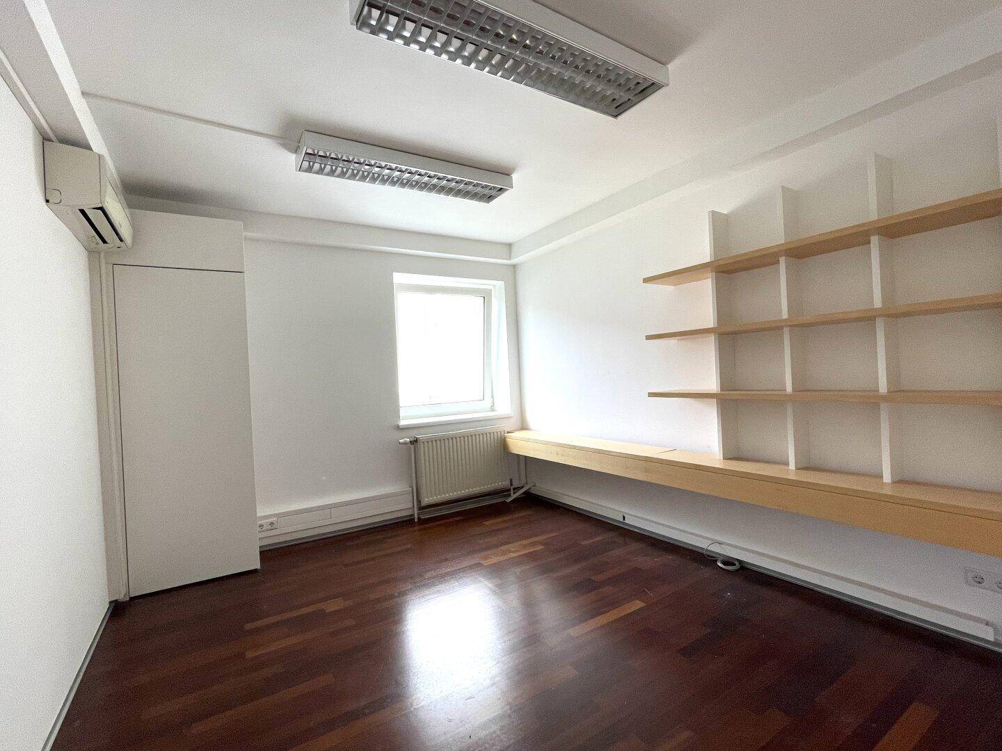 Helles Büro mit Holzboden, weißen Wänden, Fenster und eingebauten Holzregalen.