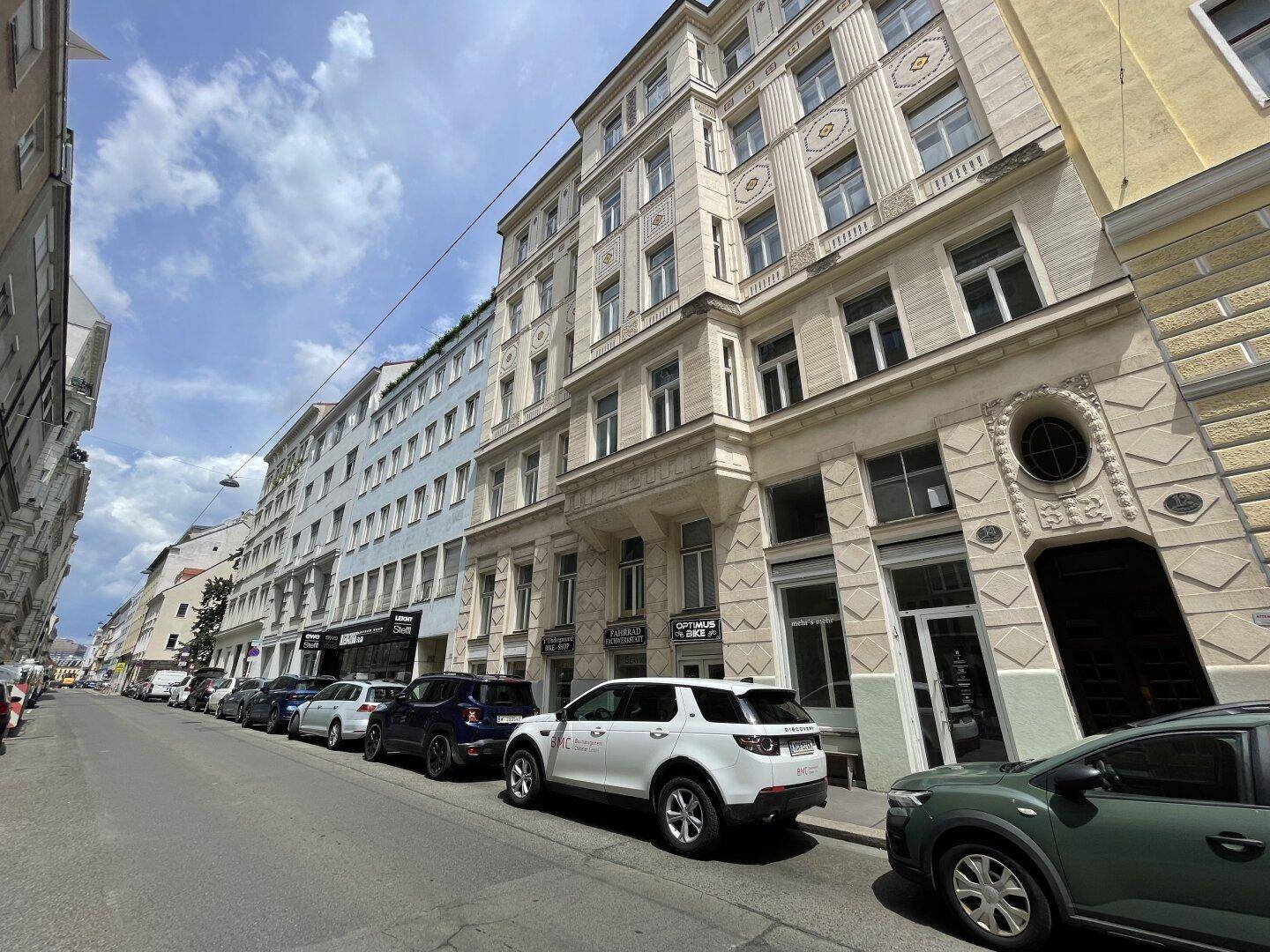 Blick auf eine belebte Stadtstraße mit parkenden Autos und mehreren historischen Wohngebäuden unter blauem Himmel.
