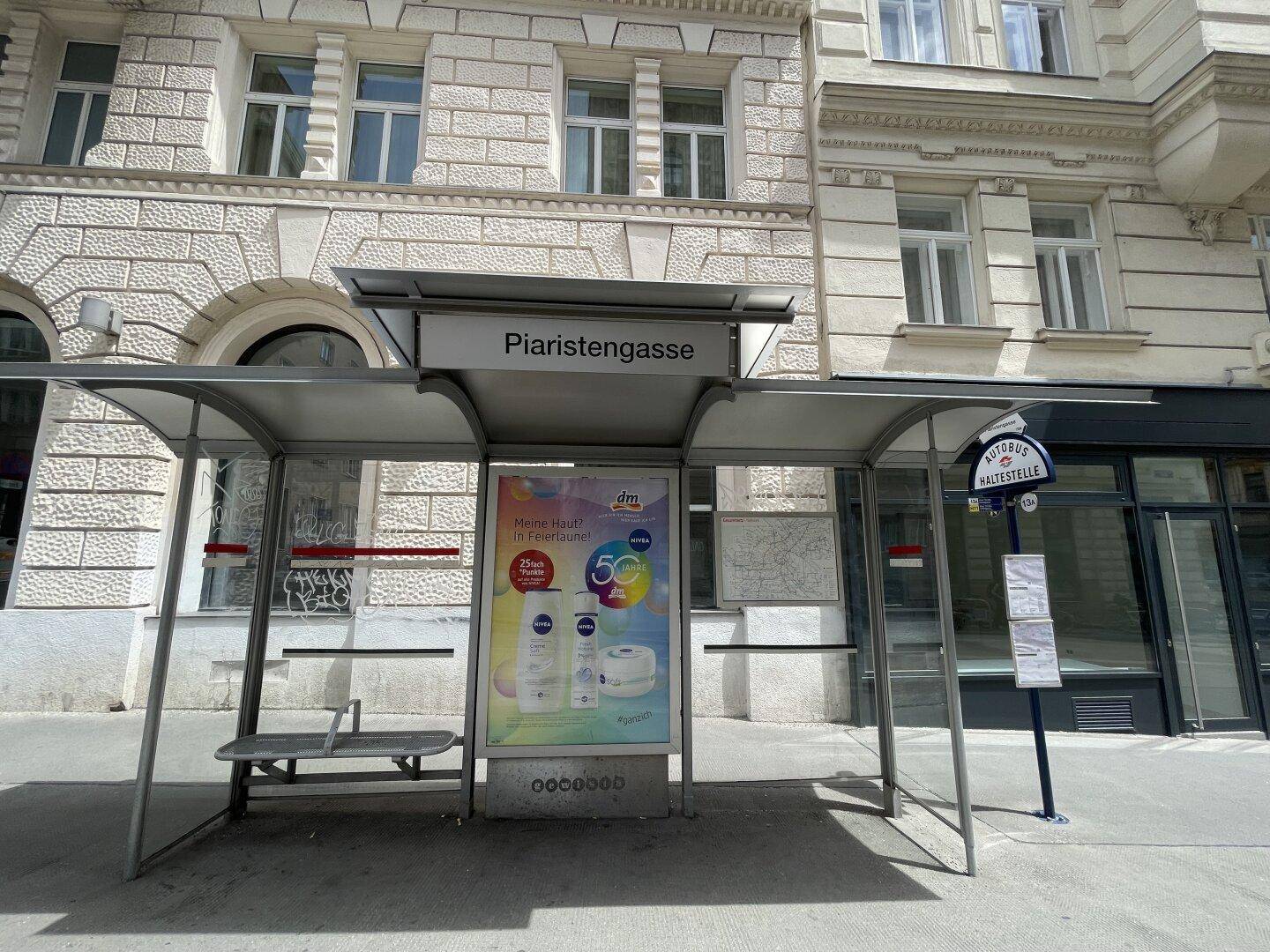 Bushaltestelle mit Werbetafel und dem Straßennamen Piaristengasse vor einer historischen Gebäudefassade.