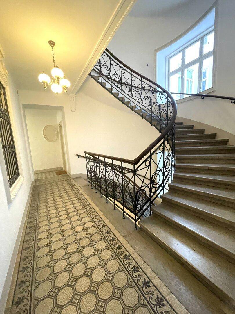 Historisches Treppenhaus mit schmiedeeisernem Geländer, gemusterten Fliesen und großen Fenstern.