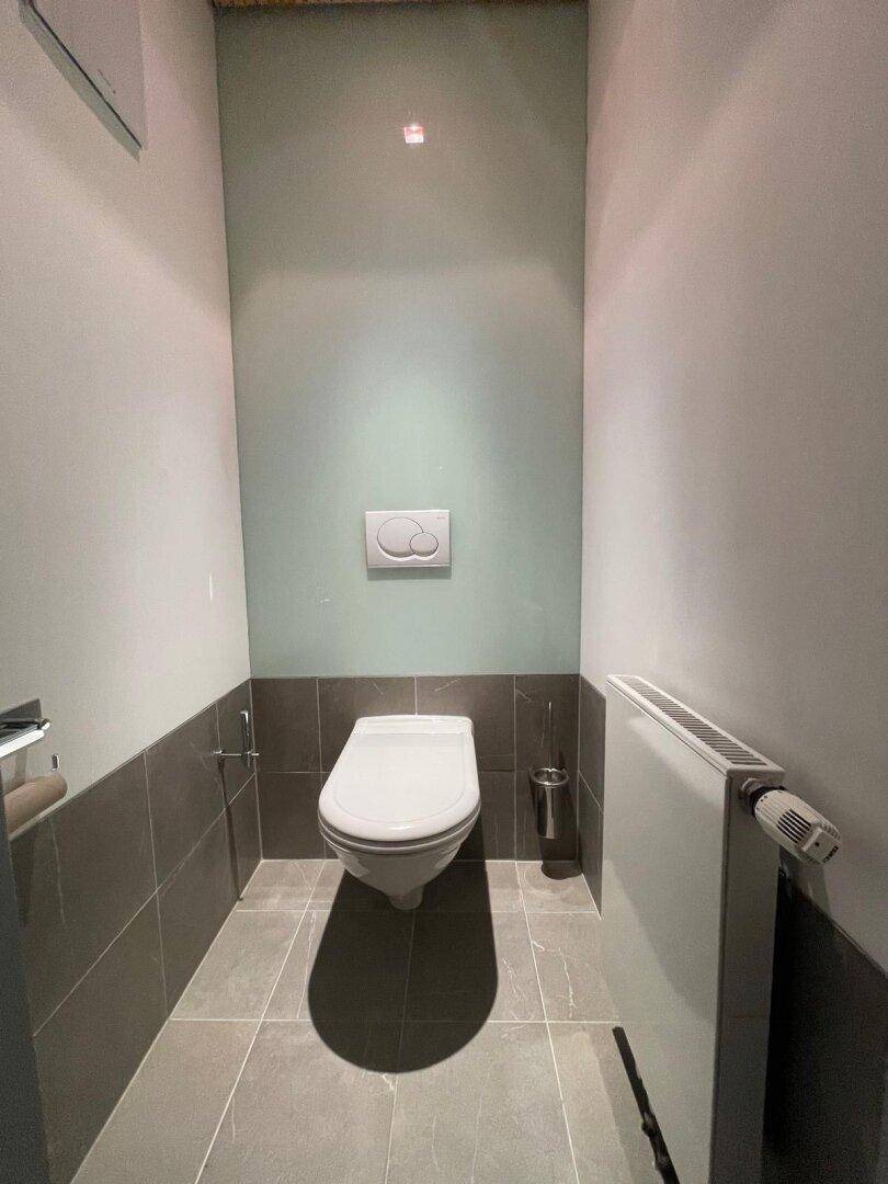 Modernes Gäste-WC mit Wand-WC, grauen Fliesen und einem Heizkörper, in einem schlichten Design.