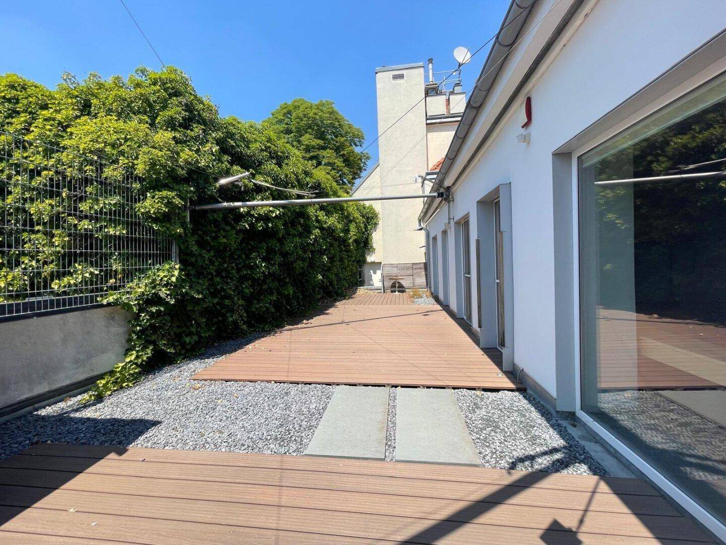 Lange Dachterrasse mit Holzdielen und Kiesflächen, umgeben von üppigem Grün und einer hohen Hecke.