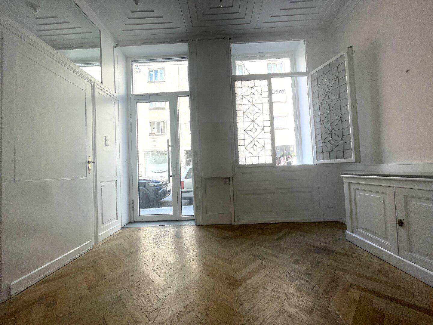 Heller Raum mit Fischgrätparkett, großen Fenstern und Zugang zur Straße.