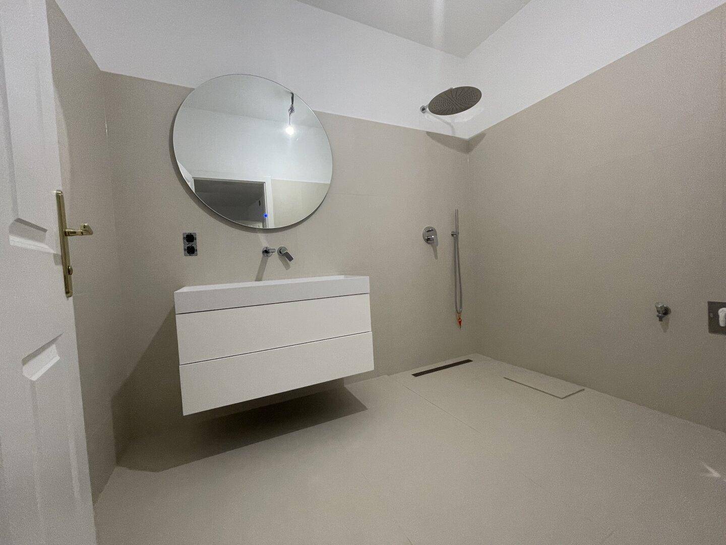 Modernes Badezimmer mit großem runden Spiegel, Waschtisch und Regendusche, in neutralen Farbtönen.
