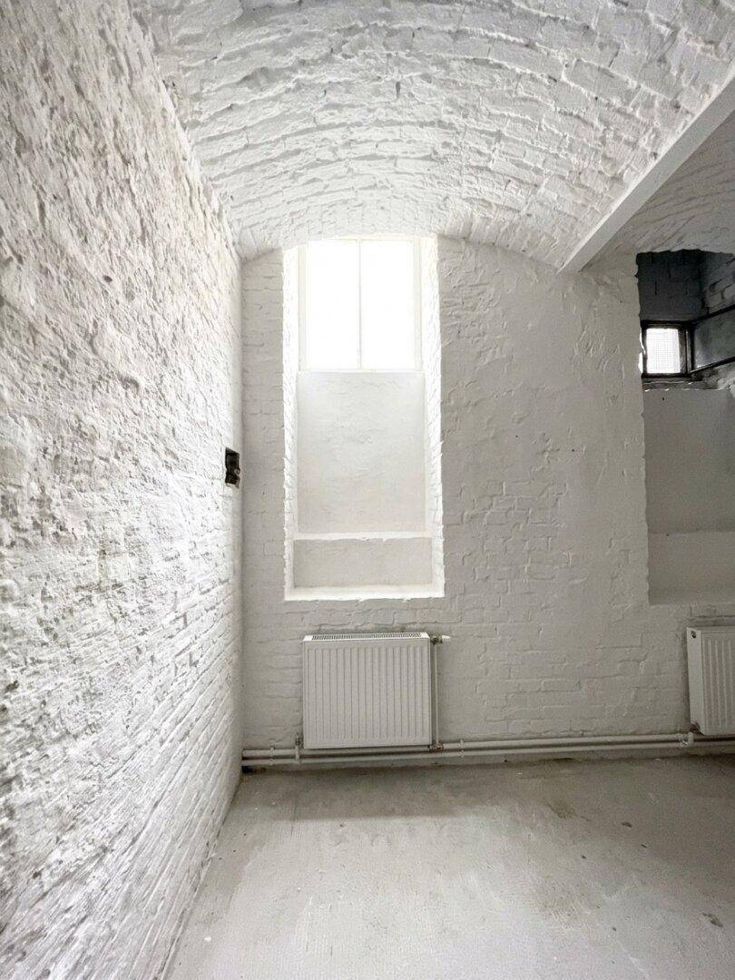 Raum mit weiß gestrichenen Ziegelwänden, Gewölbedecke, einem hohen Fenster und einem Heizkörper darunter.
