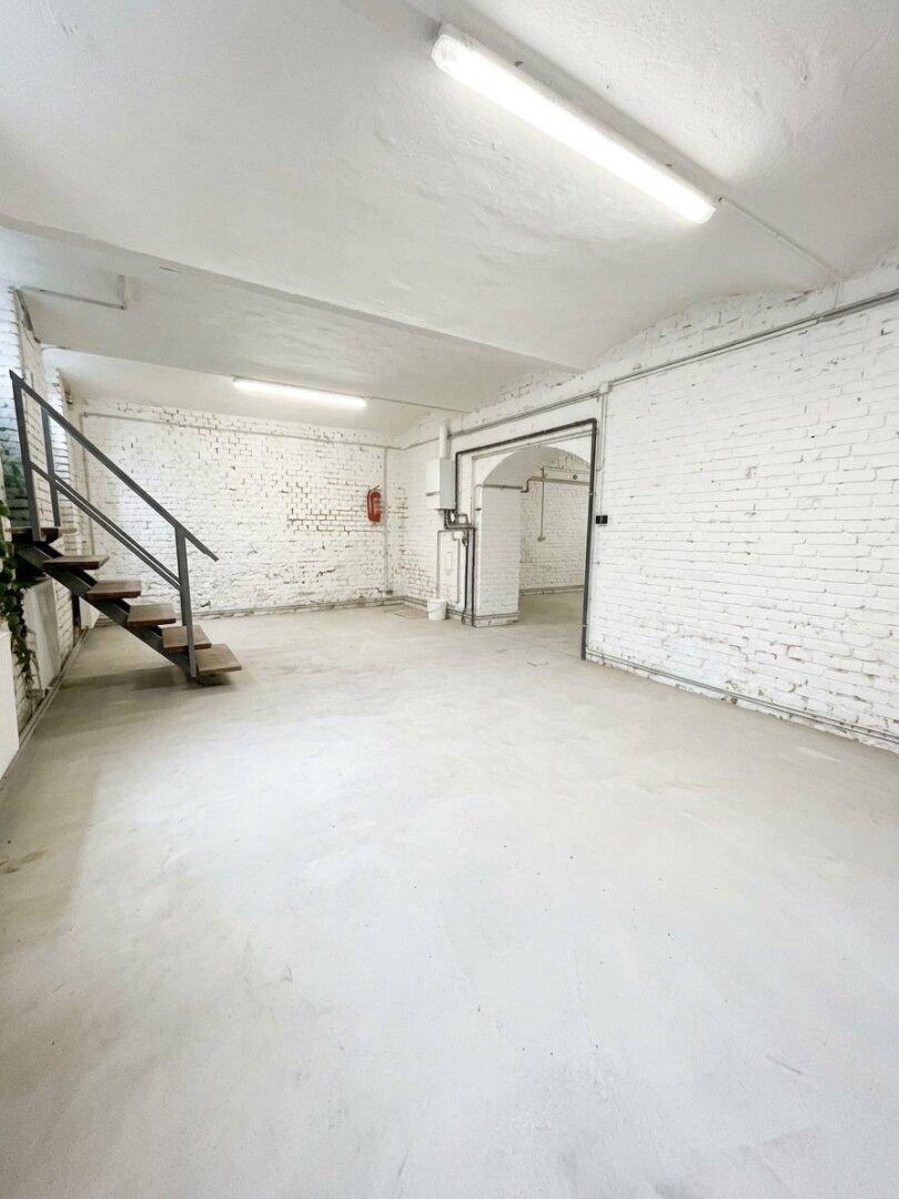 Weiterer Blick in den großen, leeren Raum mit weiß gestrichenen Ziegelwänden und sichtbaren Rohren an der Wand.