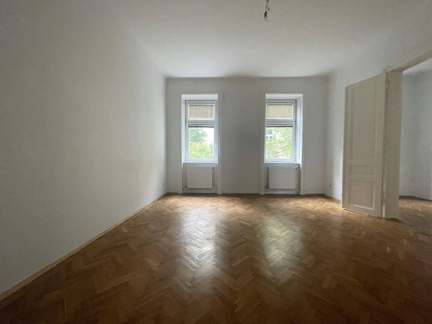 Geräumiges Zimmer mit zwei Fenstern, Fischgrätparkett und viel Tageslicht, ideal für einen Wohnbereich.