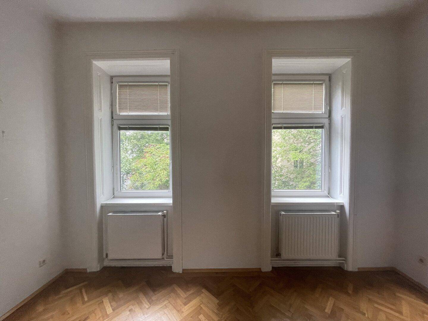 Geräumiges Zimmer mit zwei hohen Fenstern und Fischgrätparkett, das viel Licht hereinlässt.