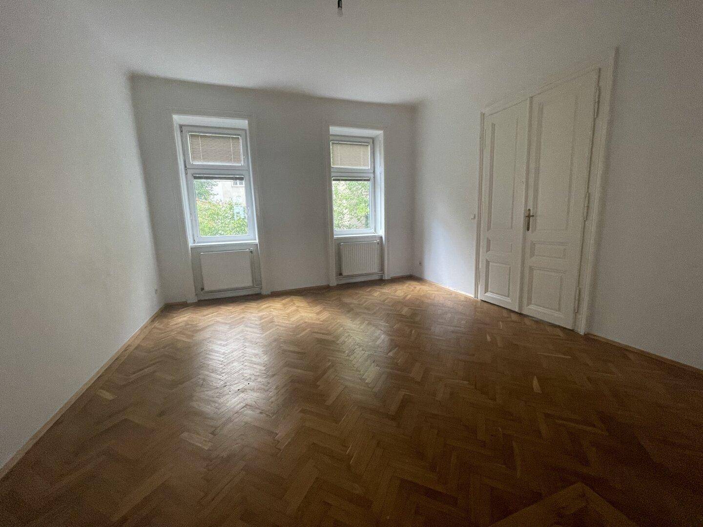 Großes Zimmer mit Fischgrätparkett und hohen Doppeltüren, ideal für einen Wohnbereich.