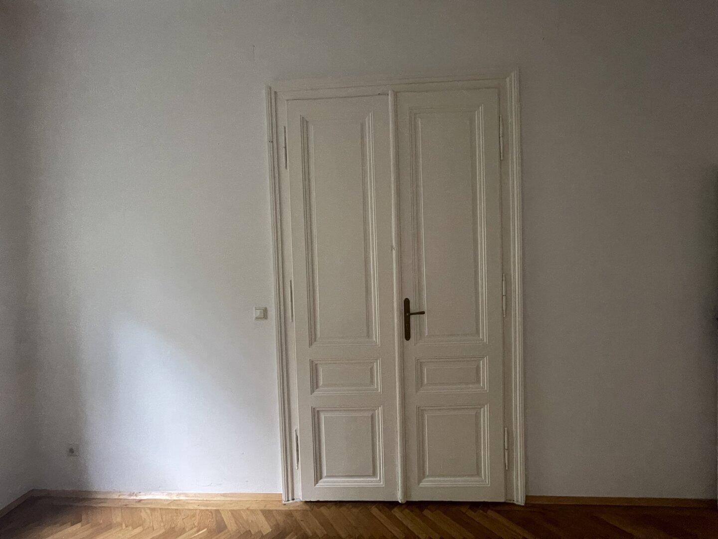 Klassische weiße Doppeltür in einem hellen Raum mit Holzboden, der zu weiteren Bereichen führt.