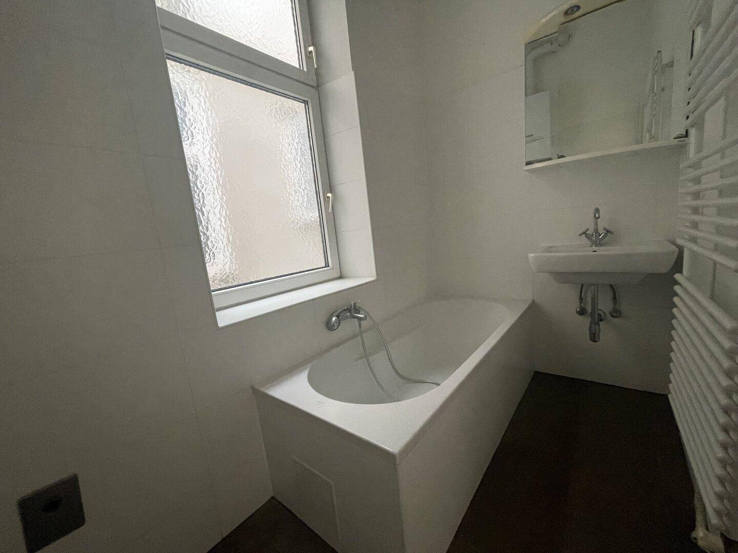 Helles Badezimmer mit Badewanne, Waschbecken und Fenster, ausgestattet mit einem Handtuchheizkörper.