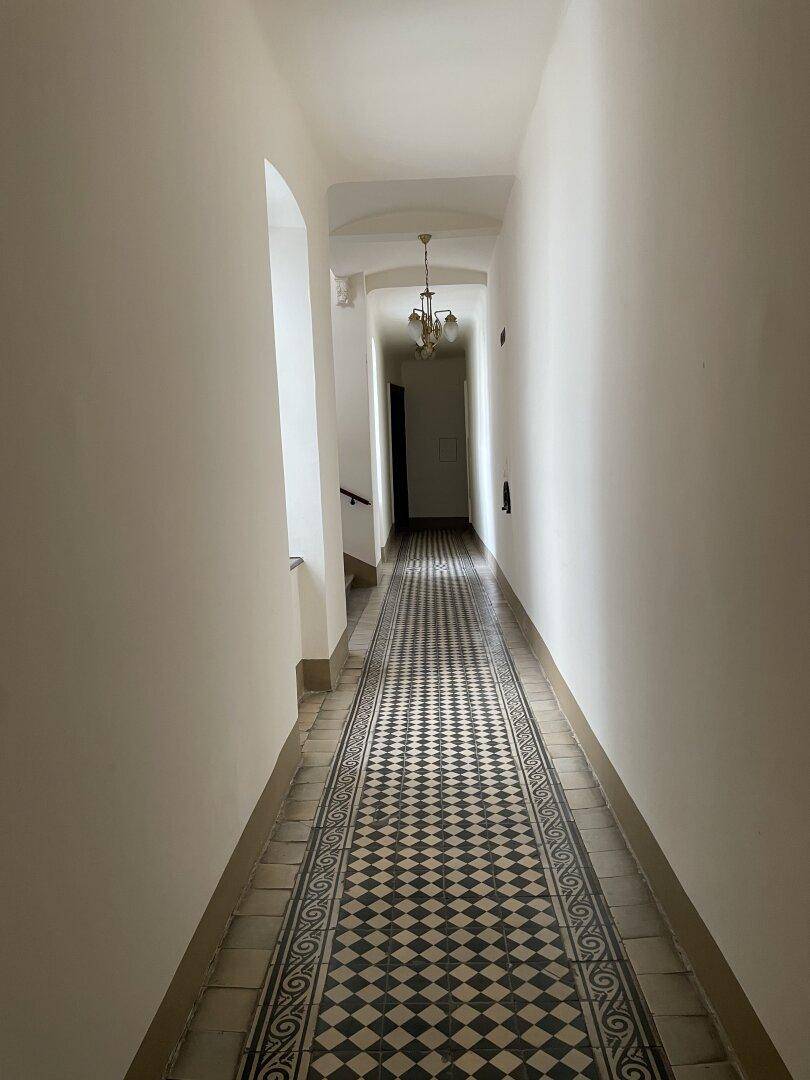 Langer, heller Flur mit gemustertem Fliesenboden und klassischem Kronleuchter, der durch das Gebäude führt.