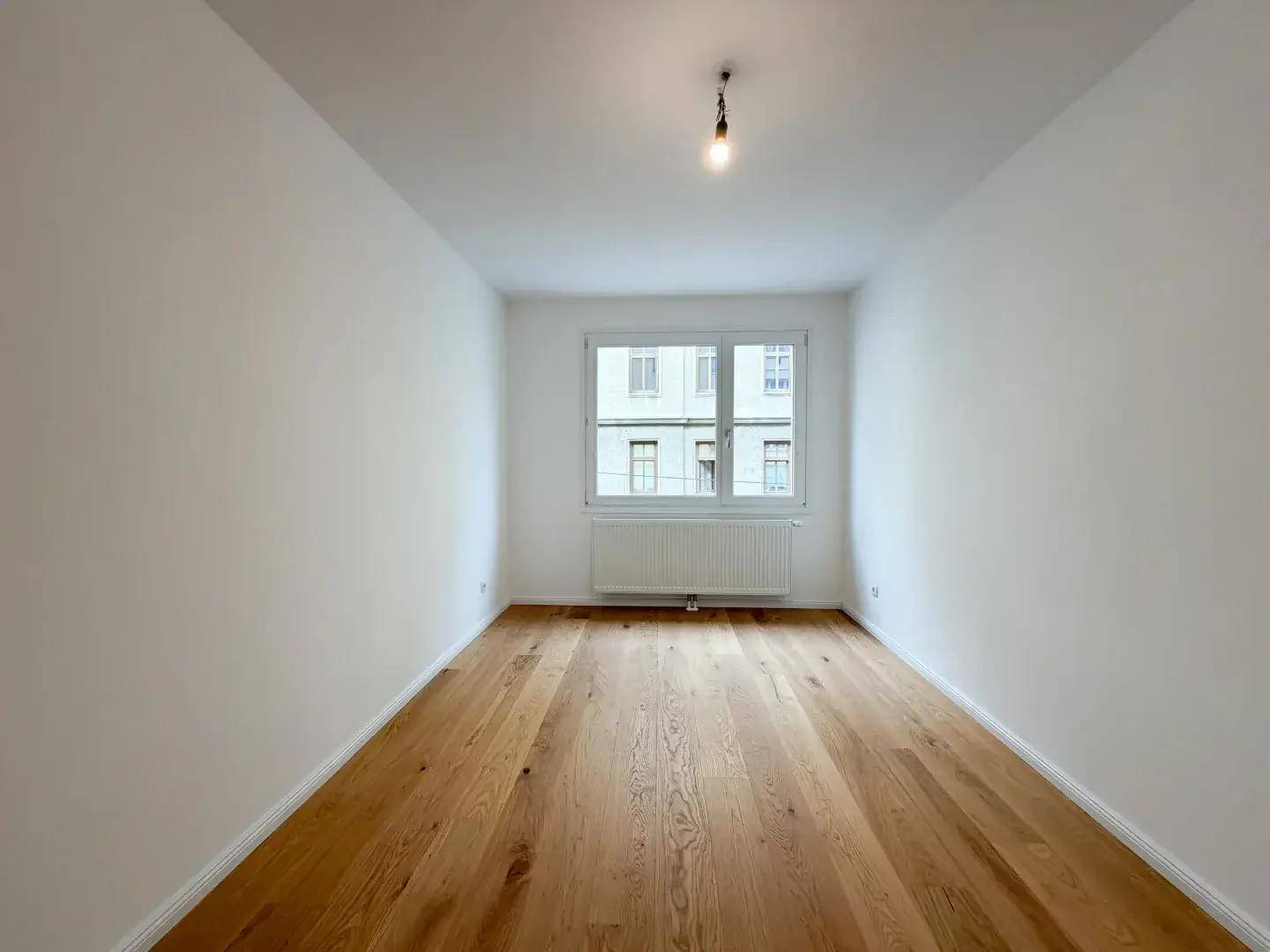 Schlichtes Schlafzimmer mit hellem Holzparkettboden und einem Fenster zur Straße.