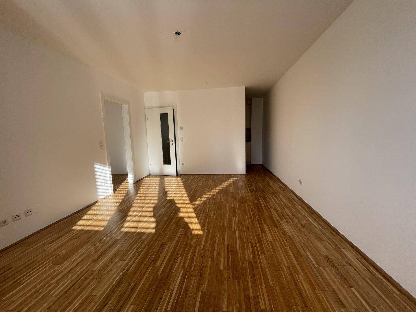 Geräumiges Zimmer mit hellem Parkettboden und weißen Wänden, durchflutet von Sonnenlicht.