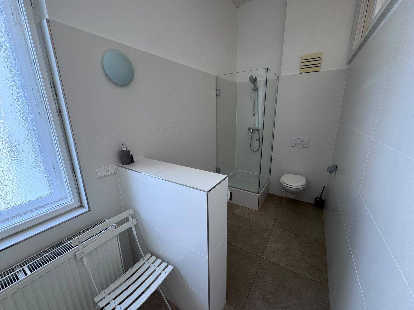 Modernes Badezimmer mit Duschkabine, WC und Fenster, hell gefliest in Weiß und Grau.