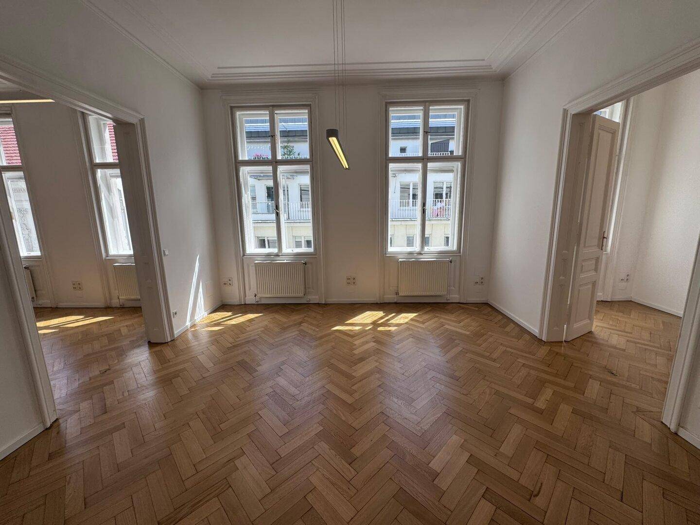 Geräumiges Wohnzimmer mit Fischgrätparkett, hohen Decken und großen Fenstern, die viel Tageslicht hereinlassen.
