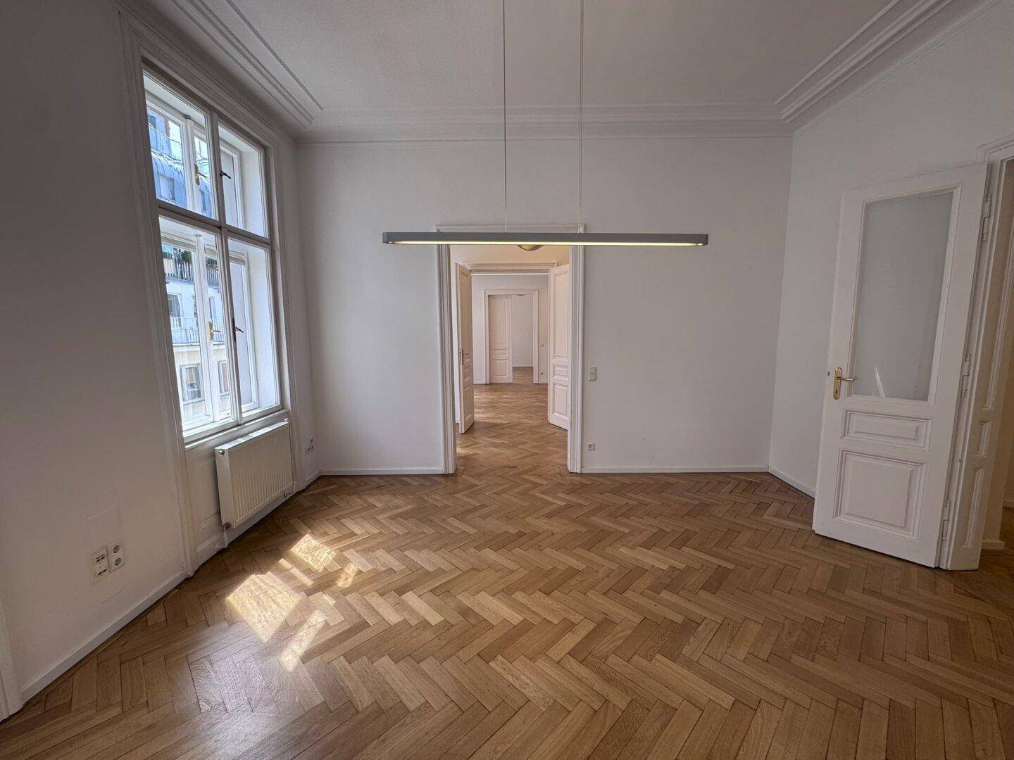 Großzügiger Raum mit Fischgrätparkett und einer modernen Deckenleuchte, der in weitere Zimmer führt.