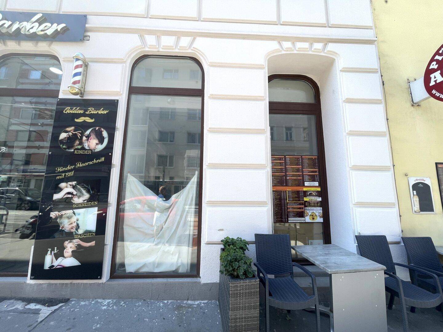 Fassade eines Gebäudes mit einem Friseursalon im Erdgeschoss und einer kleinen Außensitzgelegenheit auf dem Bürgersteig.