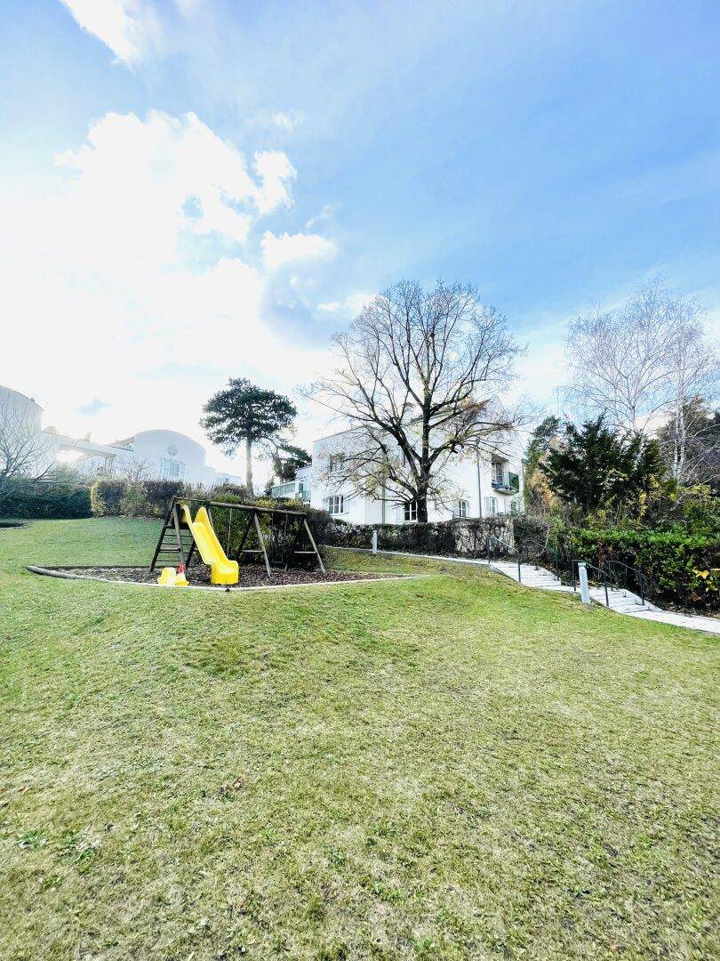 Großer Rasenbereich mit Kinderspielplatz, Rutsche und Schaukel unter blauem Himmel.