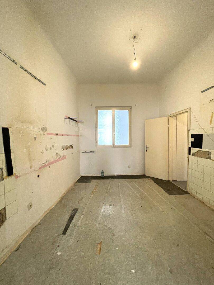 Leere Küche mit Renovierungsbedarf, Fenster und Spuren alter Installationen an den Wänden.