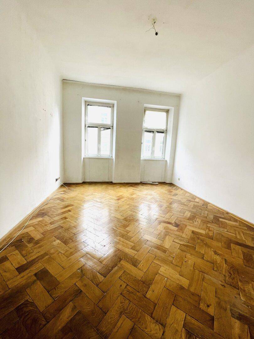Leeres Zimmer mit glänzendem Fischgrätparkett und zwei großen Fenstern, bereit zur Renovierung.