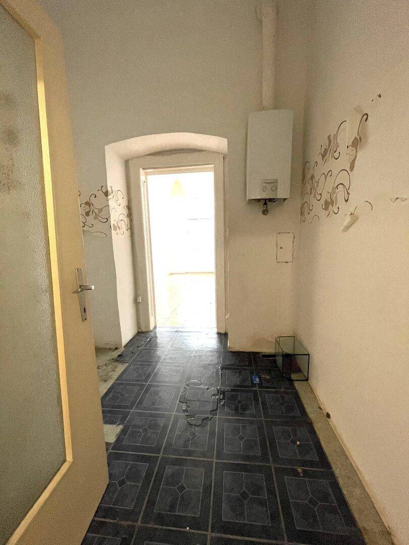 Raum mit dunklen Fliesen, einem Warmwasserbereiter und deutlichem Renovierungsbedarf.